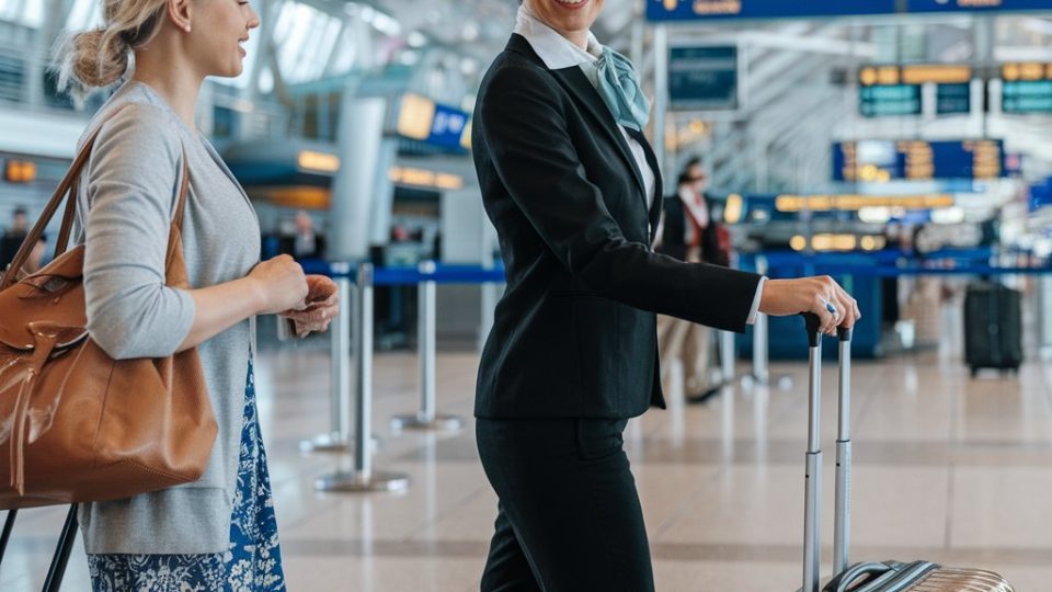 Luchthavenmedewerker helpt passagier met bagage in de terminal, met luchthavenborden en reizigers op de achtergrond.